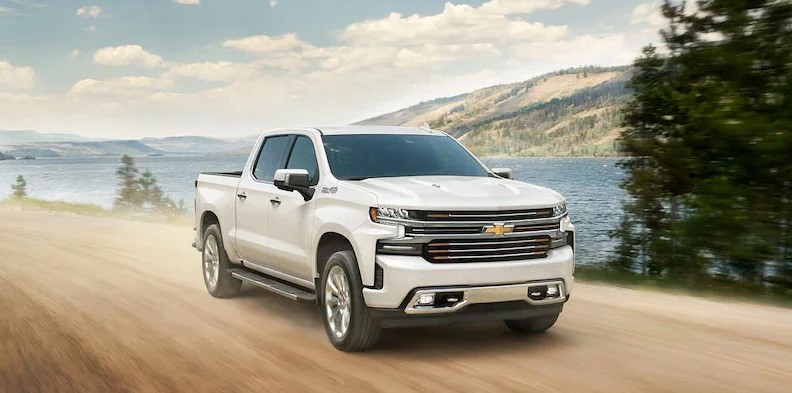 White 2021 Chevrolet Silverado 1500 driving on dirt road next to river passing by trees.