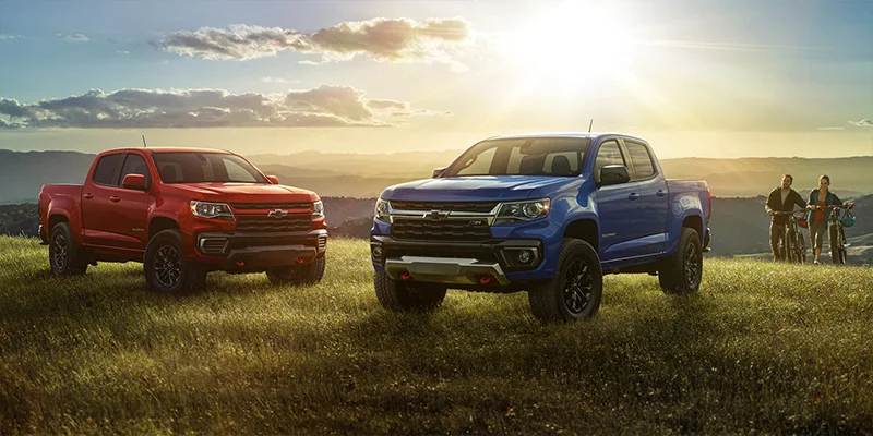 Two 2023 Chevrolet Colorados, one blue and one red, parked on top of a grassy hill with a valley in the background and two bicyclists walking up to their brand new trucks.