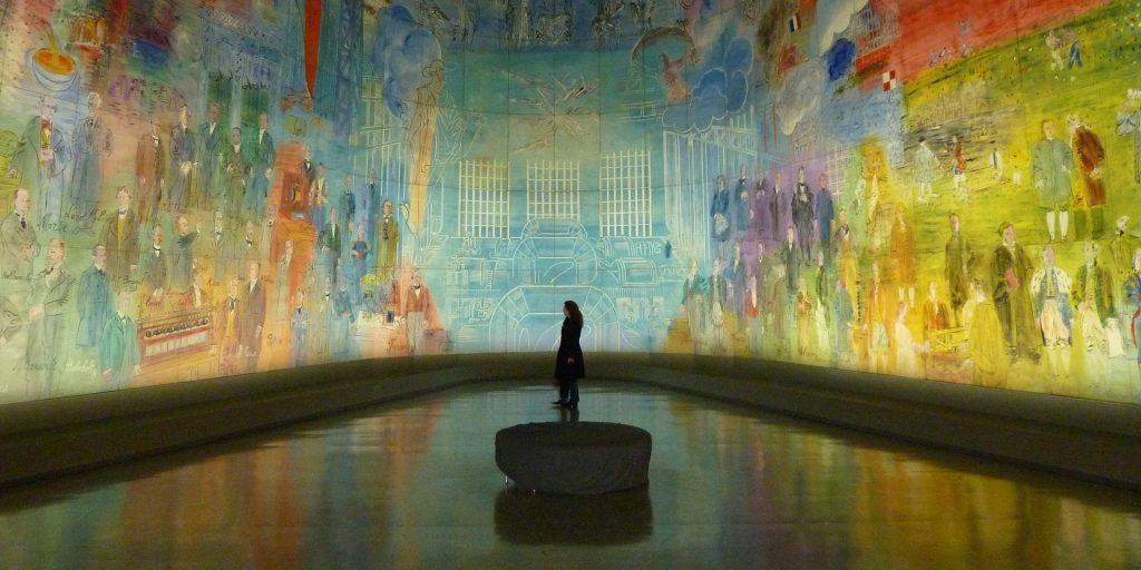 Woman standing in a large dark room with an entire panoramic art scene projected onto the wall. Similar to Van Gogh Expedition: The Immersive Experience