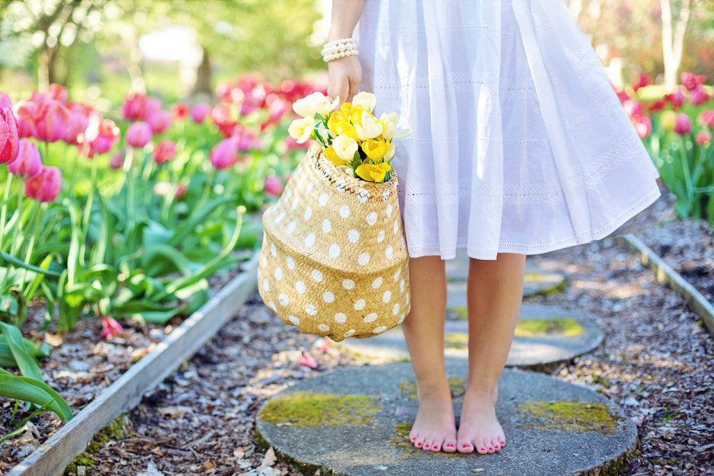 beautiful image of the bottom half of a young child wearing a white youthful dress carrying a whicker basket filled with yellow tulips. She is walking down a stone path with pink and red tulips lining the path.